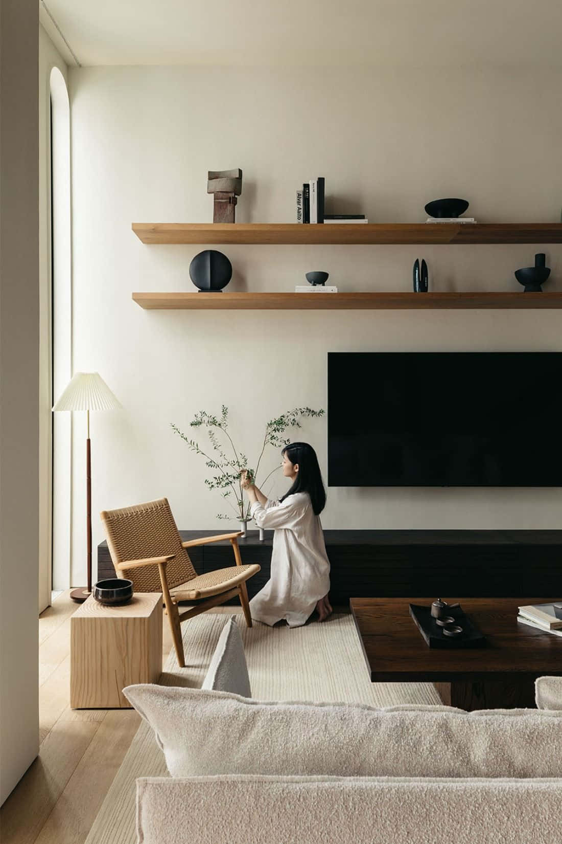 Minimalist Design 桌布 壁紙 — 一位身穿白色長袍的女性，在一個充滿柔和自然光線的簡約客廳中，將精緻的樹枝插在現代木椅旁的花瓶裡。
