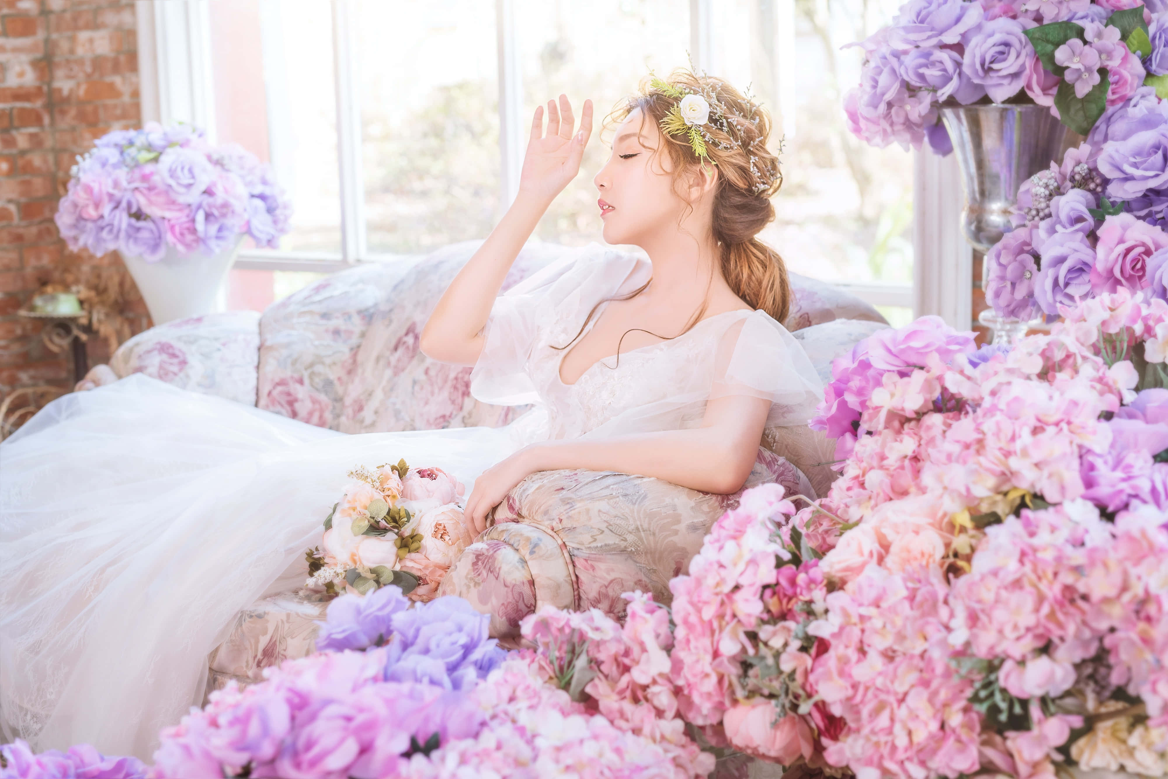 Wedding Dress 桌布 壁紙 — 一位身穿白色長裙的女性，在柔和自然光線下，優雅地坐在一張被大量粉紅色和紫色花朵環繞的花卉沙發上。