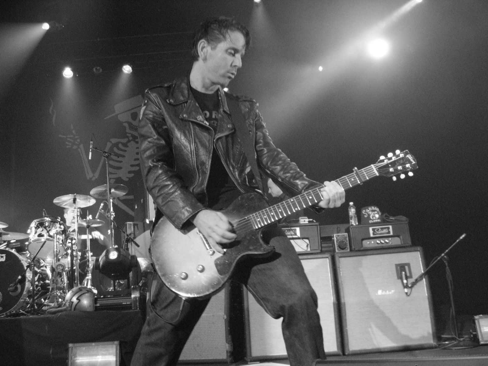 Jonny Wickersham — Social Distortion 壁紙 — 一位身穿皮褸的音樂家在燈光昏暗的舞台上熱情地彈奏電結他，身後有擴音器和鼓。