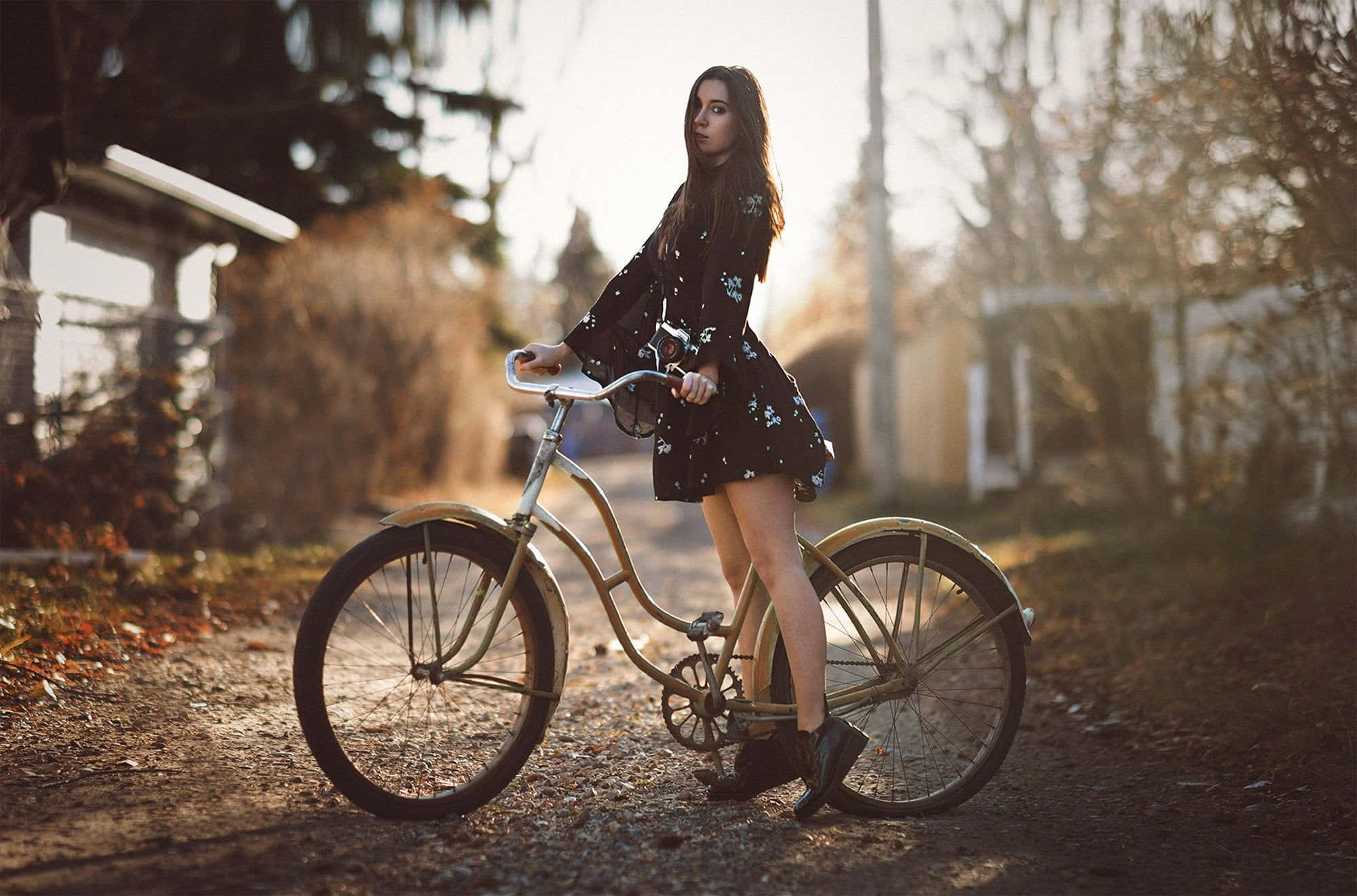 Beach Cruiser 桌布 壁紙 — 一位身穿碎花裙的年輕女子站在一條陽光明媚的泥濘小徑上，旁邊是一輛復古單車，四周環繞著秋天的樹木。