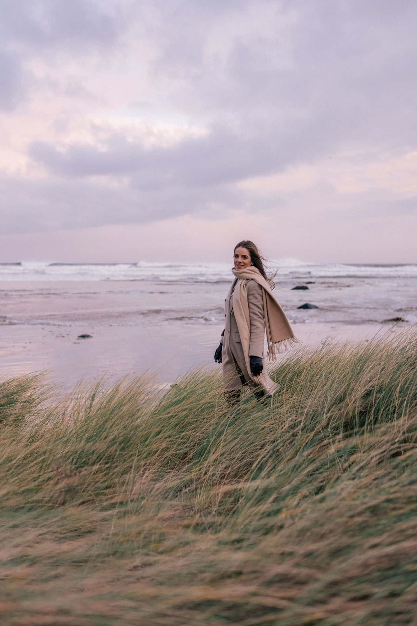 Winter Beach 桌布 壁紙 — 一位身穿米色外套和圍巾的女性，在黃昏時分站在風吹沙灘的高草叢中微笑，天空呈現柔和的紫色和粉紅色。