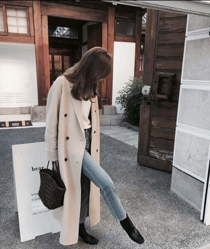Winter Fashion 桌布 壁紙 — 一位身穿米色外套和牛仔褲的女性靠在一棟帶有木質裝飾和石徑的現代建築上。
