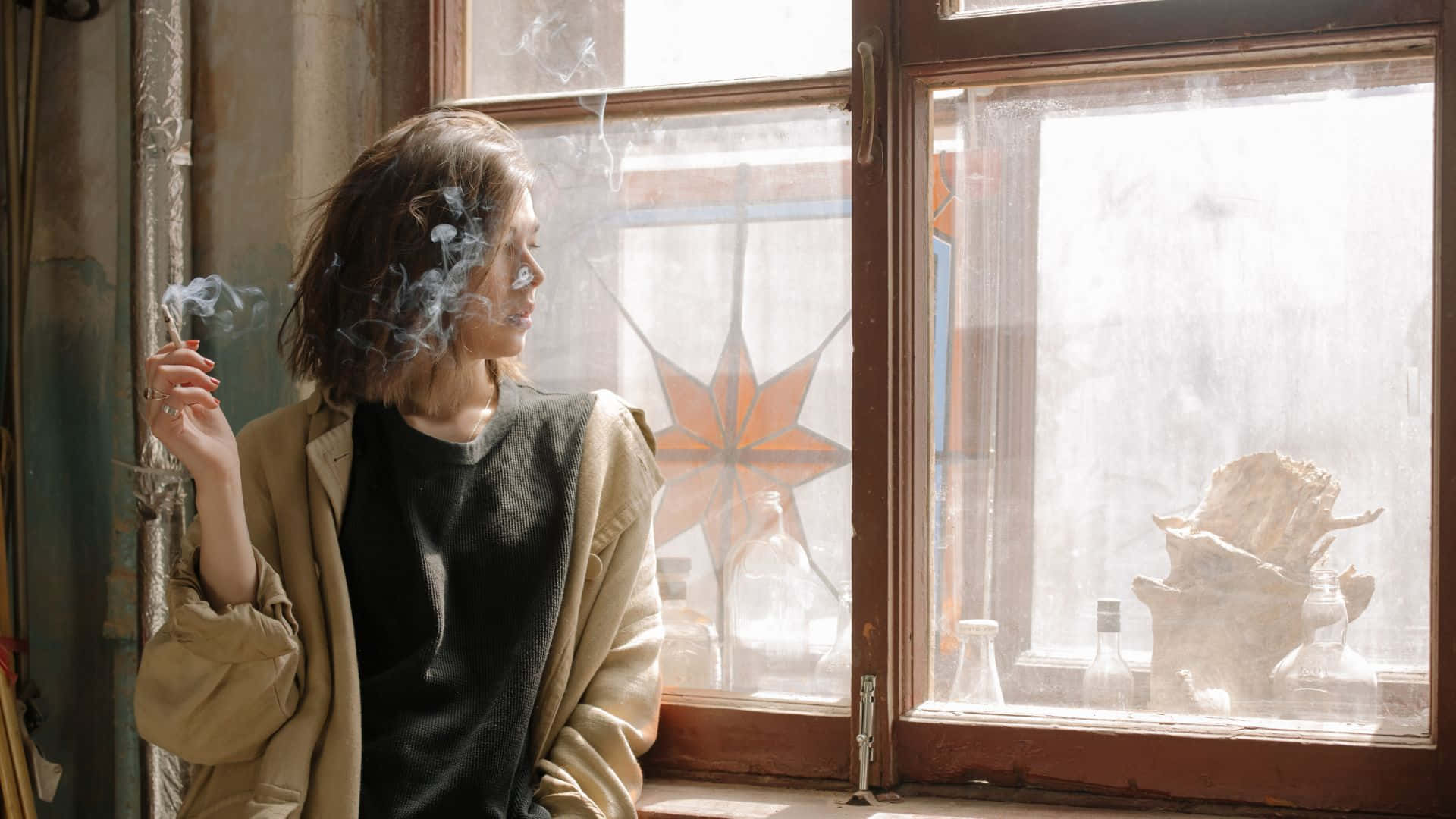吸煙 桌布 壁紙 — 一位身穿米色外套的女性站在佈滿灰塵的窗邊，呼出煙霧，陽光透過彩色玻璃星星照射進來。