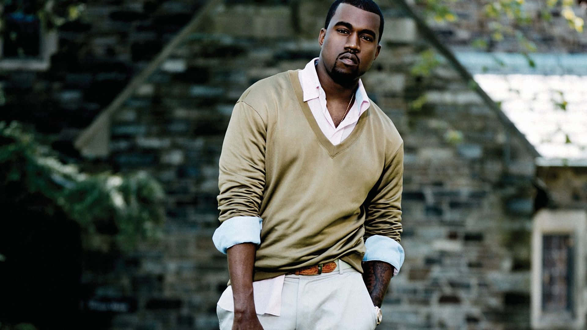 Kanye West 桌布 壁紙 — 一位身穿米色毛衣和白色褲子的男子自信地站在有綠色樹葉的石牆旁。