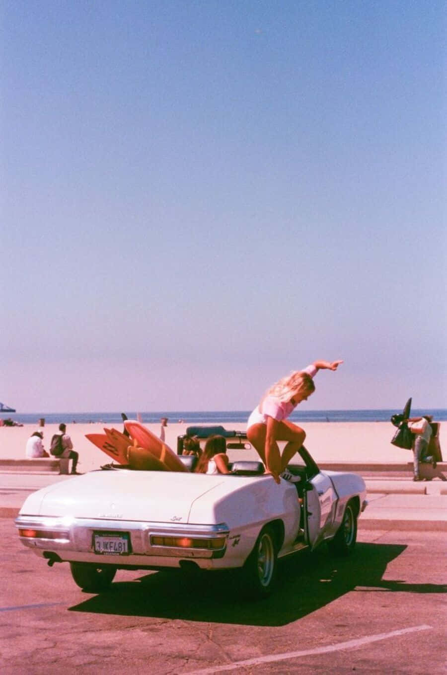 90's Vintage Retro Aesthetic 桌布 壁紙 — 一位身穿粉紅色泳衣的女性在停泊於陽光明媚的海灘上的白色開篷車上擺姿勢，背景可見衝浪板和海洋。