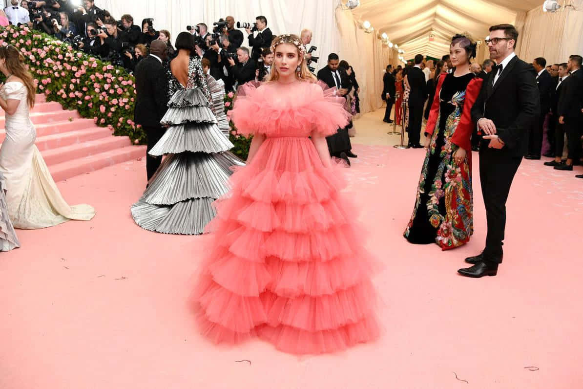 Met Gala 桌布 壁紙 — 一位身穿粉紅色蓬鬆薄紗長裙的女性，在一個華麗的晚會上走過粉紅色地毯，周圍有攝影師和穿著華麗服飾的賓客。