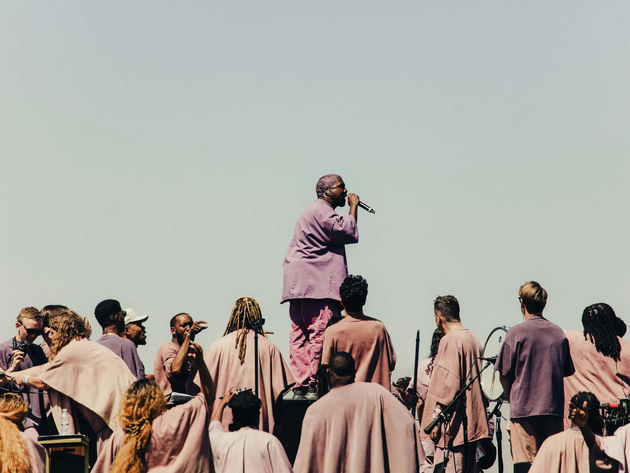 Kanye 桌布 壁紙 — 一位身穿粉紅色西裝的表演者在舞台上對著麥克風唱歌，而一群穿著相配粉紅色長袍的人群在蒼白的天空下觀看。