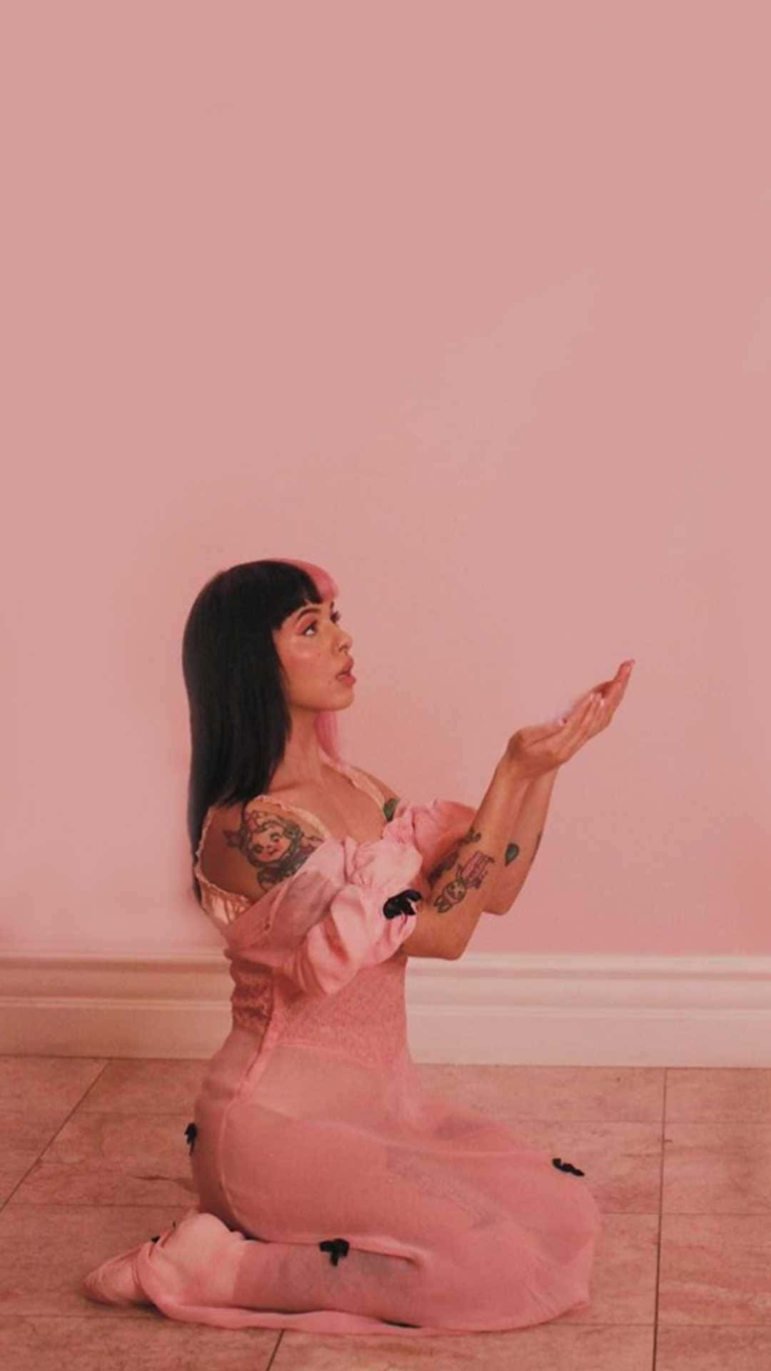 Melanie Martinez 桌布 壁紙 — 一位身穿粉紅色連衣裙的女性跪在瓷磚地板上，背靠粉紅色牆壁，手臂上的紋身清晰可見。