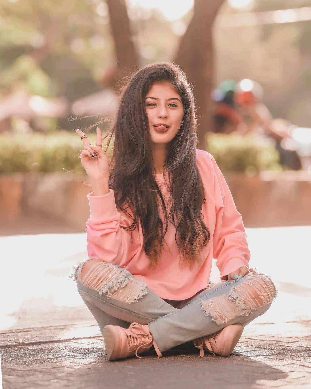 Pink Sweater 桌布 壁紙 — 一位身穿粉紅色運動衫和破洞牛仔褲的年輕女子盤腿坐在人行道上，頑皮地伸出舌頭並比出和平手勢。