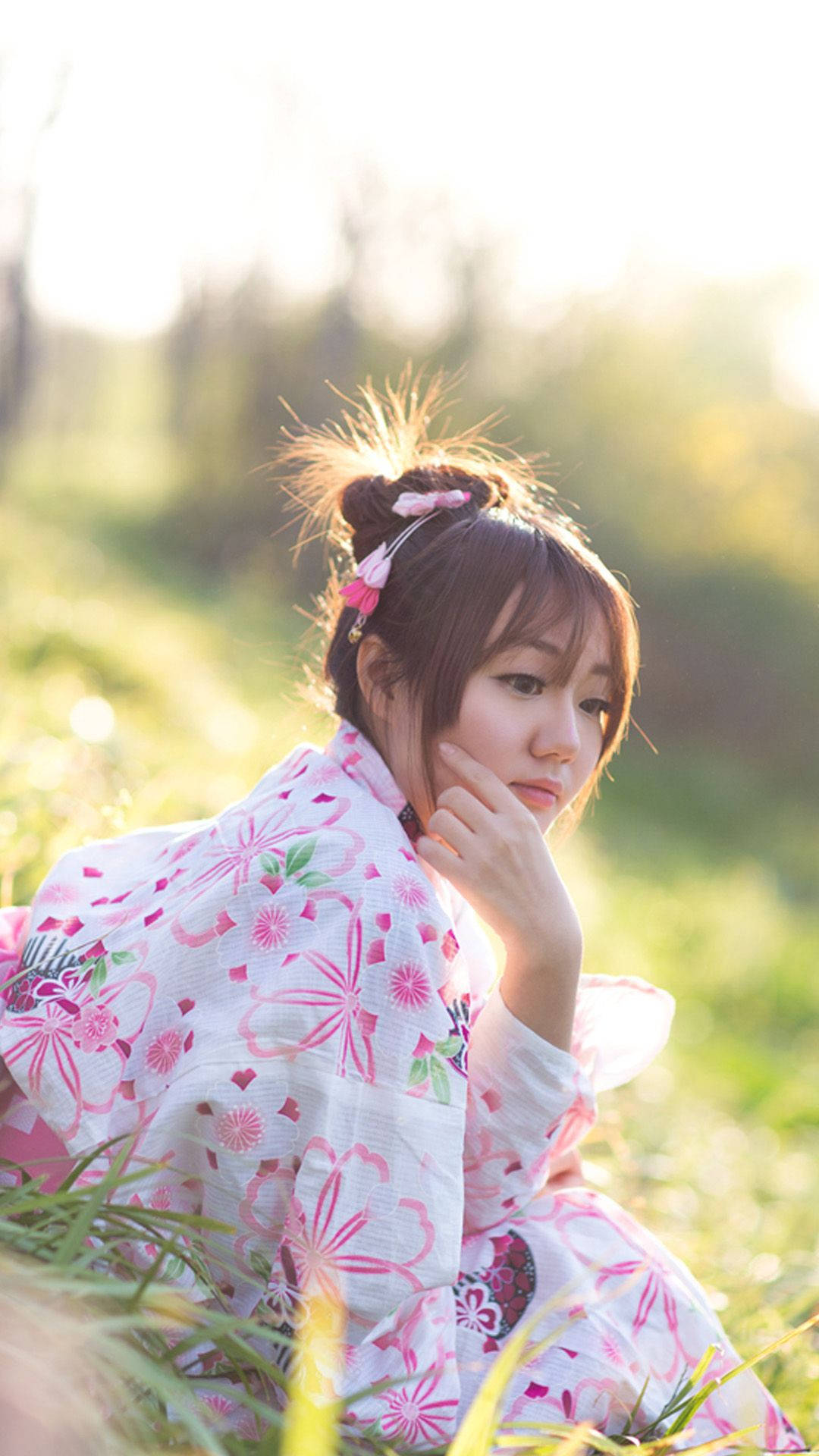 Beautiful Girl 桌布 壁紙 — 一位身穿粉紅花卉和服的年輕女子坐在陽光普照的田野中，她的手輕輕觸碰臉頰，金色的光線從她身後的草叢中透出。
