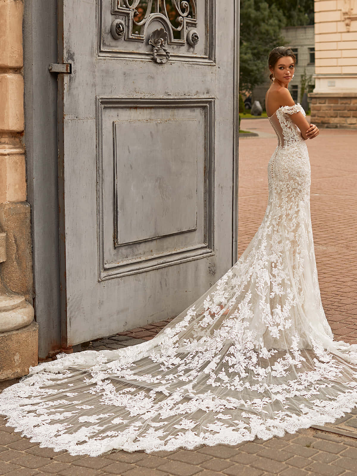 Bride's Dress 桌布 壁紙 — 一位身穿精緻白色蕾絲禮服的新娘，在陽光明媚的磚砌庭院中，優雅地站在一扇風化的灰色門旁。