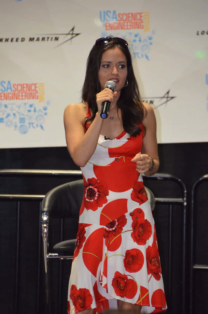 Danica Mckellar 桌布 壁紙 — 一位身穿紅白罌粟花裙的女性在科學工程活動中對著麥克風講話。