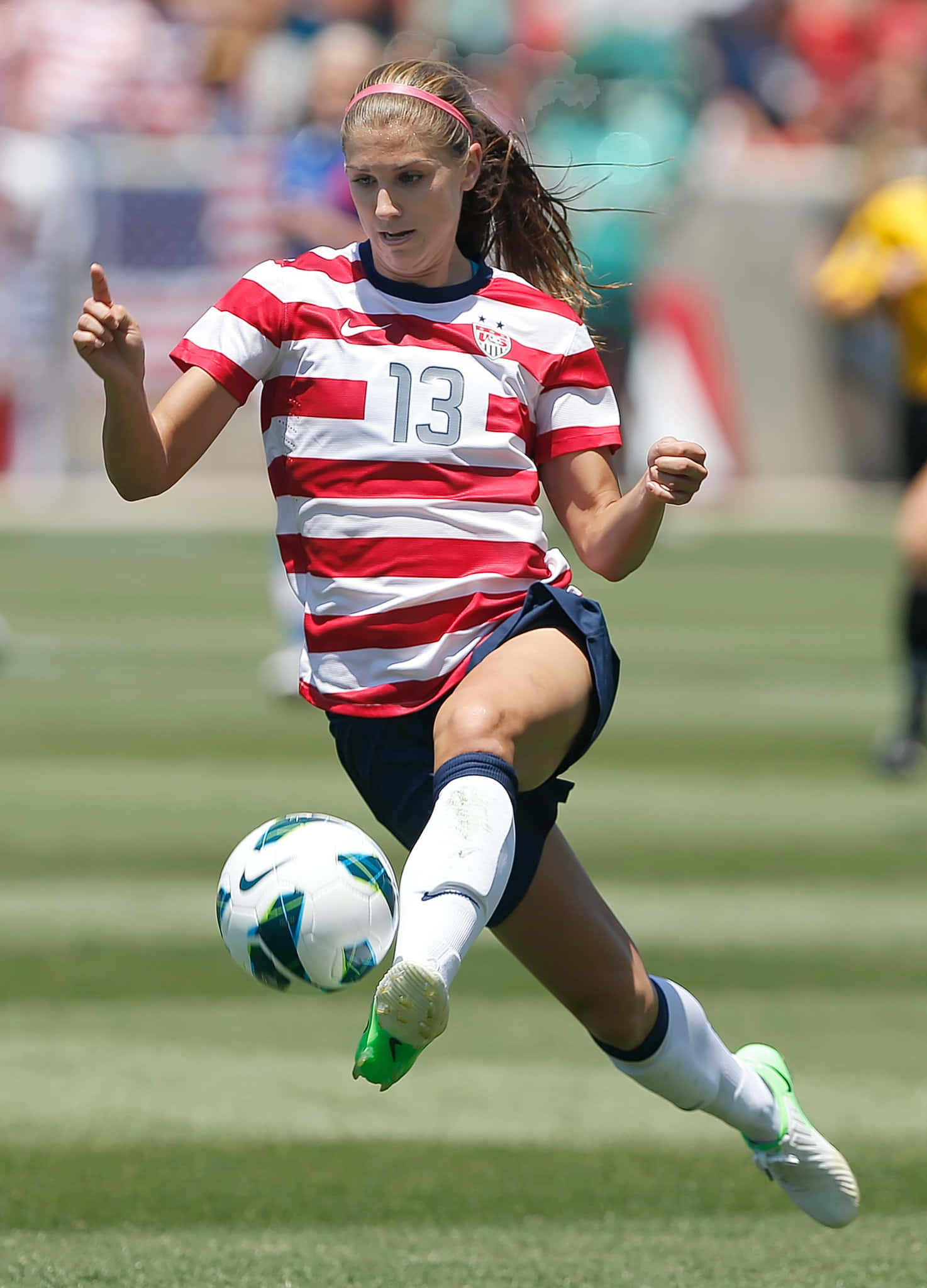 Alex Morgan 桌布 壁紙 — 一位身穿紅白間條球衣13號的女足球員在陽光明媚的綠色球場上凌空踢球。