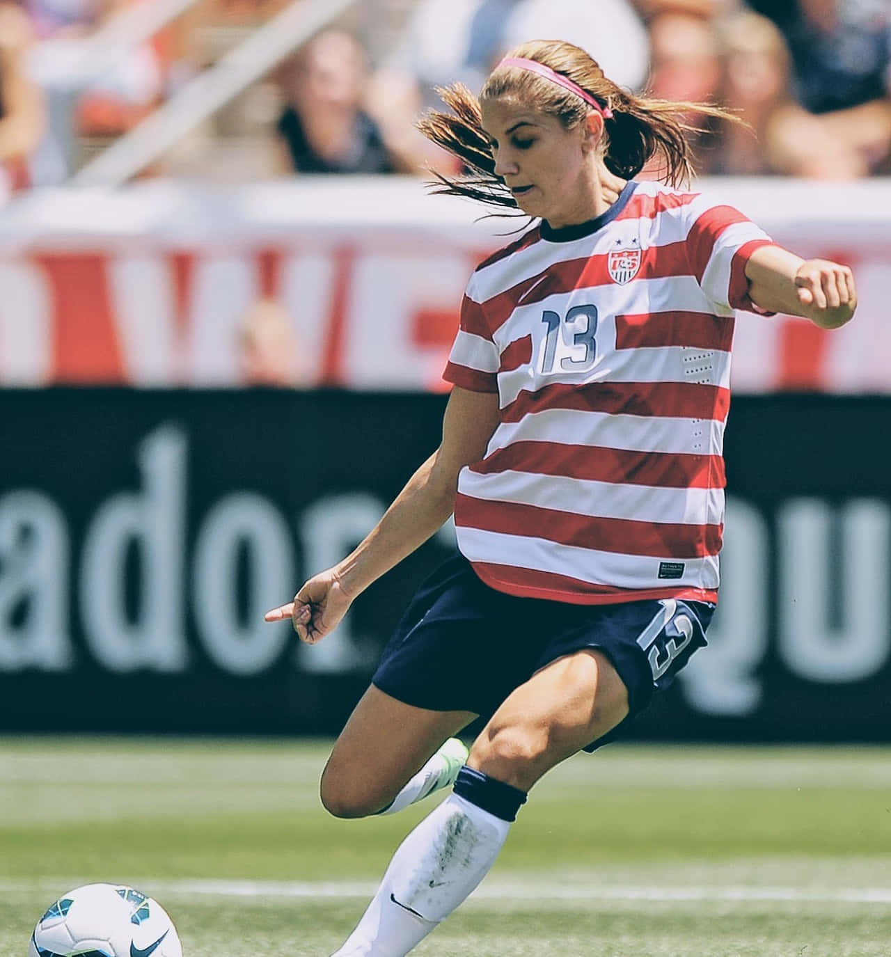 Alex Morgan 桌布 壁紙 — 一位身穿紅白間條球衣的女足球員在陽光明媚的球場上踢球，背景有模糊的觀眾。