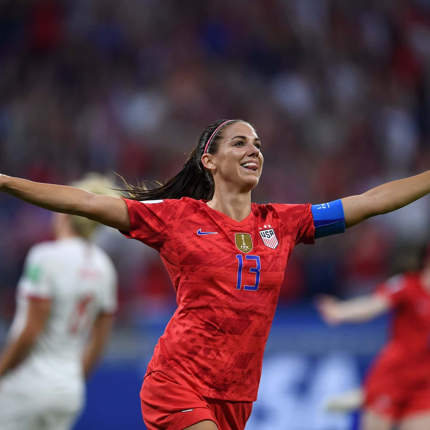 Alex Morgan 桌布 壁紙 — 一位身穿紅色13號球衣的美國女足球員在球場上張開雙臂勝利慶祝。