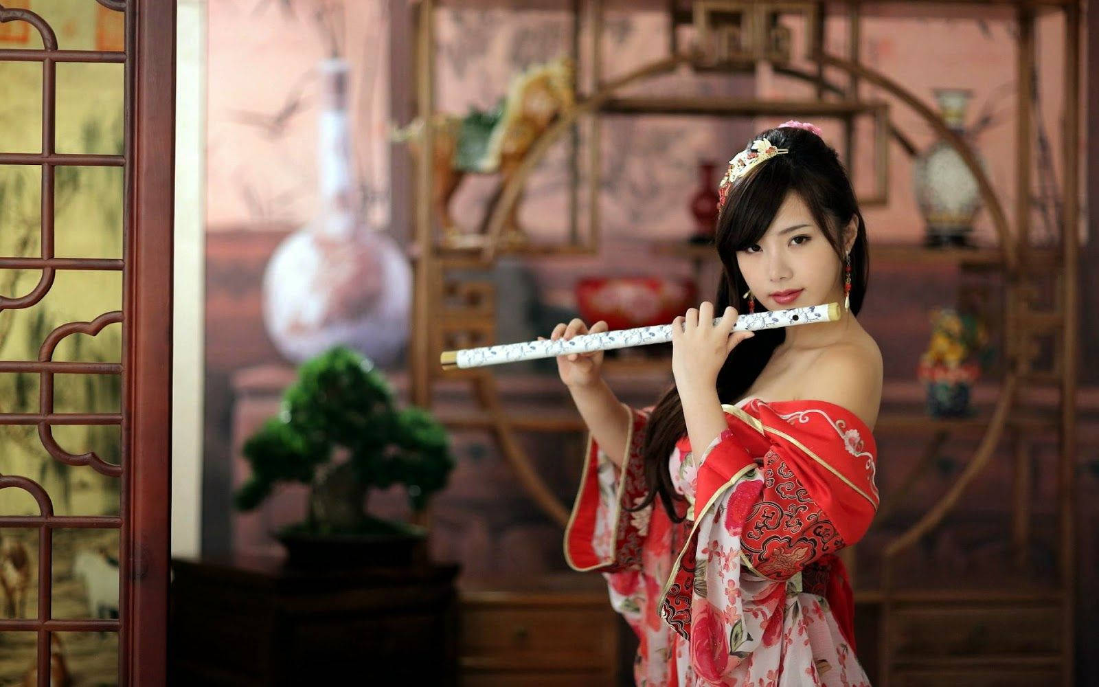  壁紙 — 一位身穿紅色傳統長袍的女性在一個帶有裝飾架和盆景的木製室內吹奏長笛。