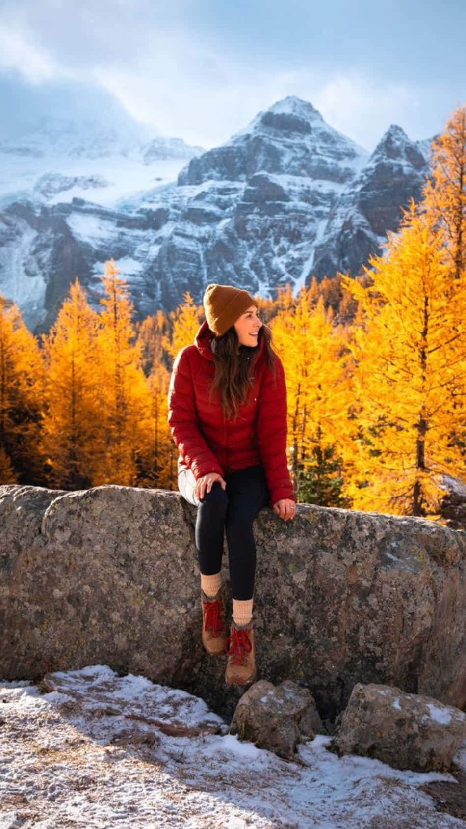 Fall Hikes 桌布 壁紙 — 一位身穿紅色外套的女性坐在岩石上，微笑著望向被金色秋樹環繞的雪山。