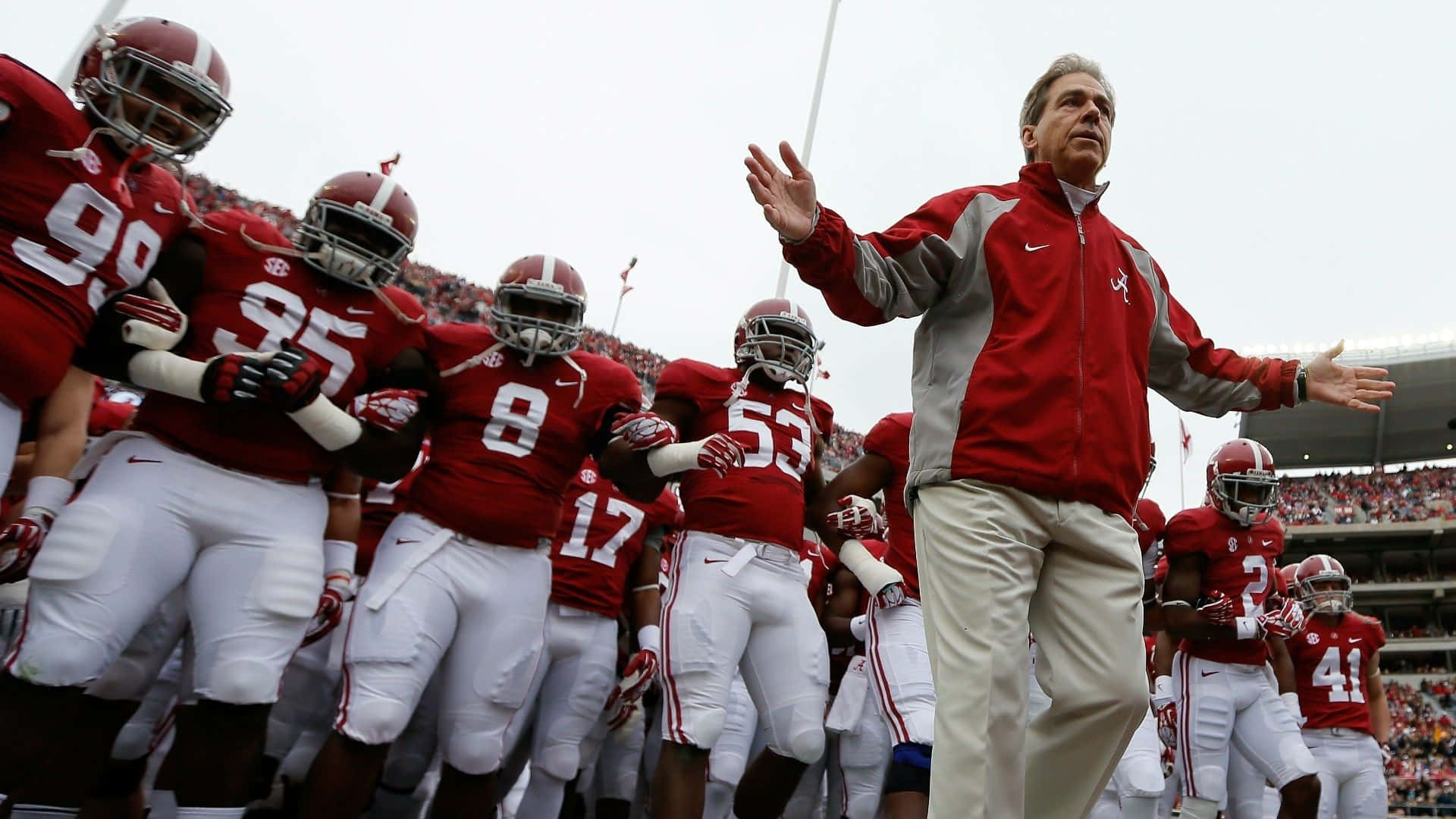 「Roll Tide Roll!」 壁紙 — 一位身穿紅色外套的足球教練在比賽中熱情地向場上身穿紅白球衣的球隊做出手勢。