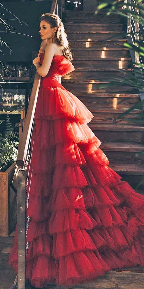Red Wedding 桌布 壁紙 — 一位身穿紅色多層長裙的女性，優雅地站在木樓梯旁，旁邊有綠植。