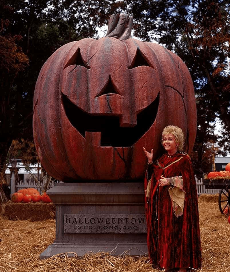 Halloweentown 桌布 壁紙 — 一位身穿紅色天鵝絨服裝的女性微笑地站在一個刻有Halloween Town字樣的巨型雕刻南瓜雕像旁。