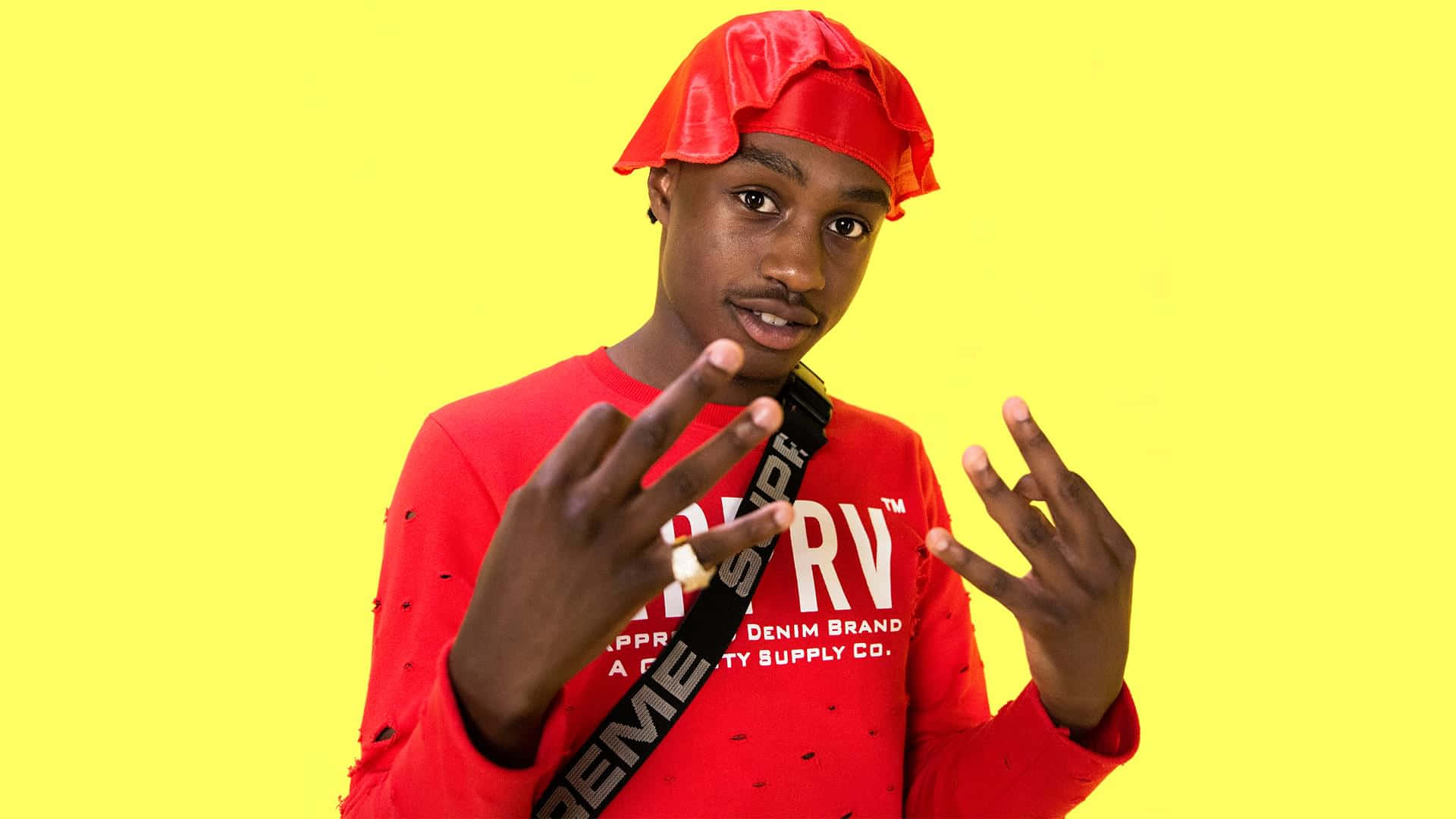 Lil Tjay 桌布 壁紙 — 一位身穿紅色服裝和配套帽子的年輕男子在明亮的黃色背景前自信地擺姿勢。