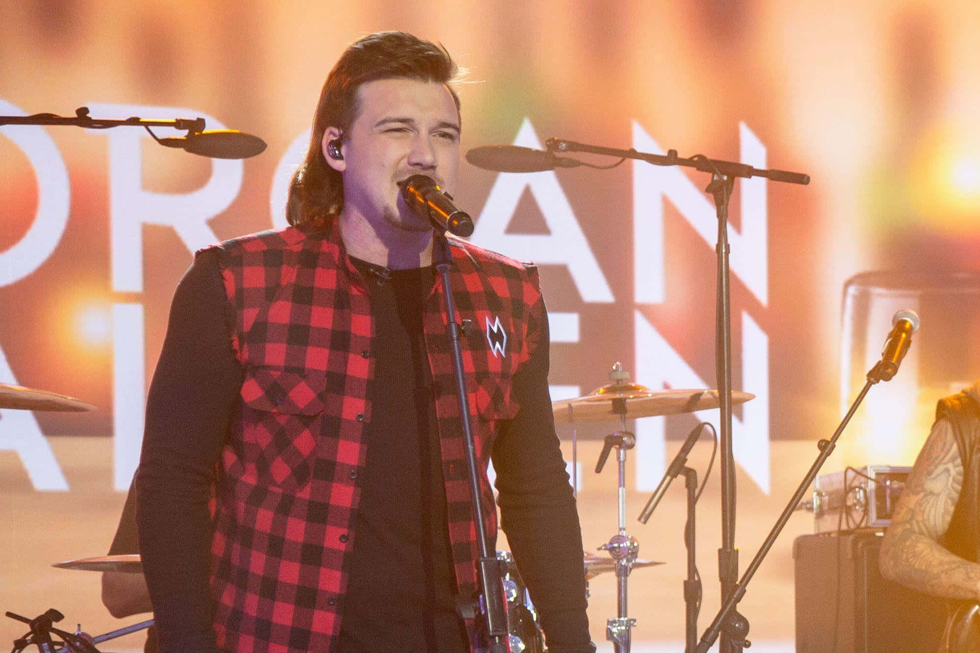 Morgan Wallen 桌布 壁紙 — 一位身穿紅色格仔背心的男歌手在溫暖燈光的舞台上，對著咪高峰熱情演唱，背景有鼓組和發光字母。