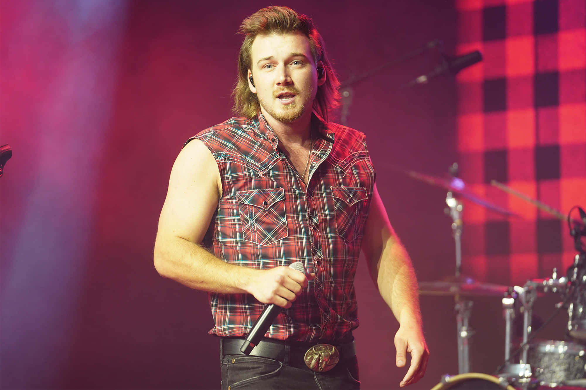 Morgan Wallen 桌布 壁紙 — 一位身穿紅色格仔背心的男歌手在舞台上表演，背景有鼓組，舞台燈光為鮮豔的粉紅色和紫色。