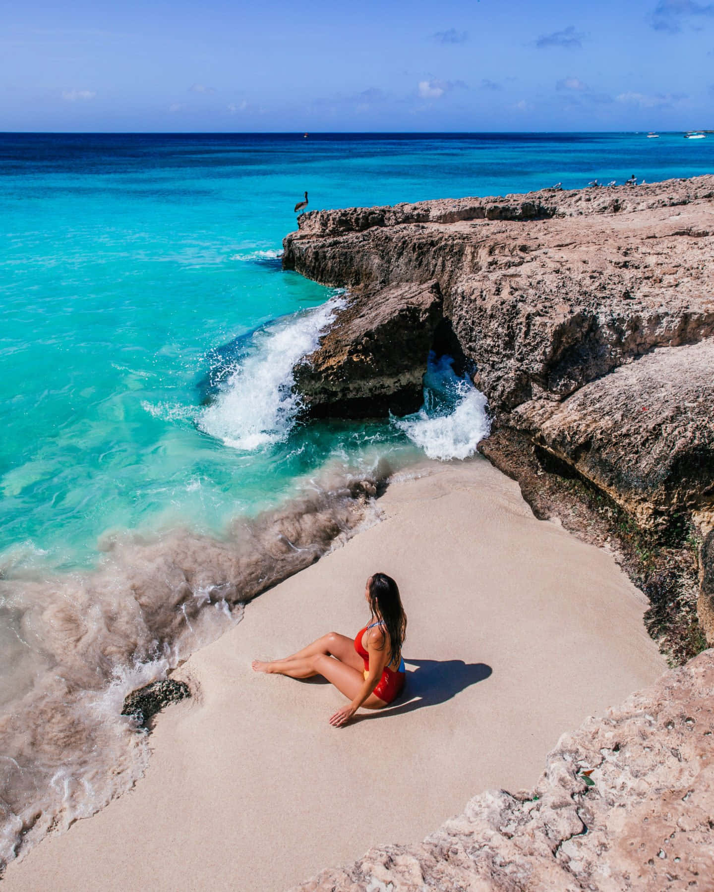 Aruba 桌布 壁紙 — 一位身穿紅色泳衣的女性平靜地坐在沙灘上，在清澈的藍天下，碧綠的海浪拍打著崎嶇的懸崖。