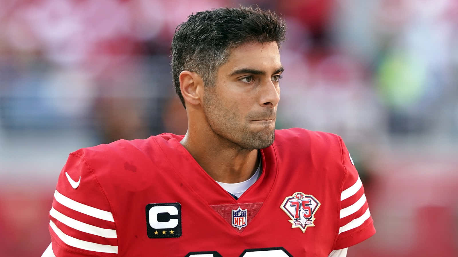 Jimmy Garoppolo 桌布 壁紙 — 一位身穿紅色球衣、帶有隊長臂章的美式足球員，在日間比賽中專注地凝視。