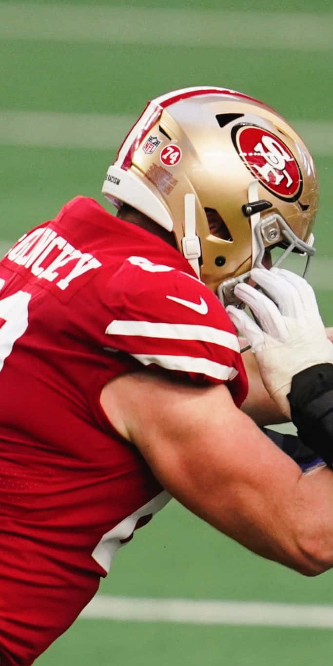 Mike Mcglinchey 桌布 壁紙 — 一位身穿紅色球衣和金色頭盔的三藩市49人美式足球員在綠色球場上調整頭盔。