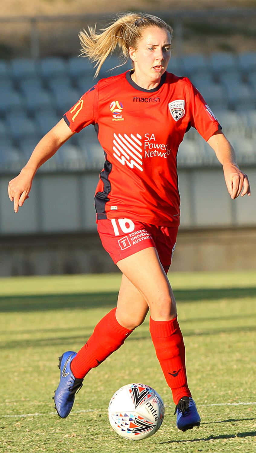 Adelaide United 桌布 壁紙 — 一位身穿紅色球衣的女足球員在陽光照耀的草地上運著一個白色足球，身後是空無一人的看台。