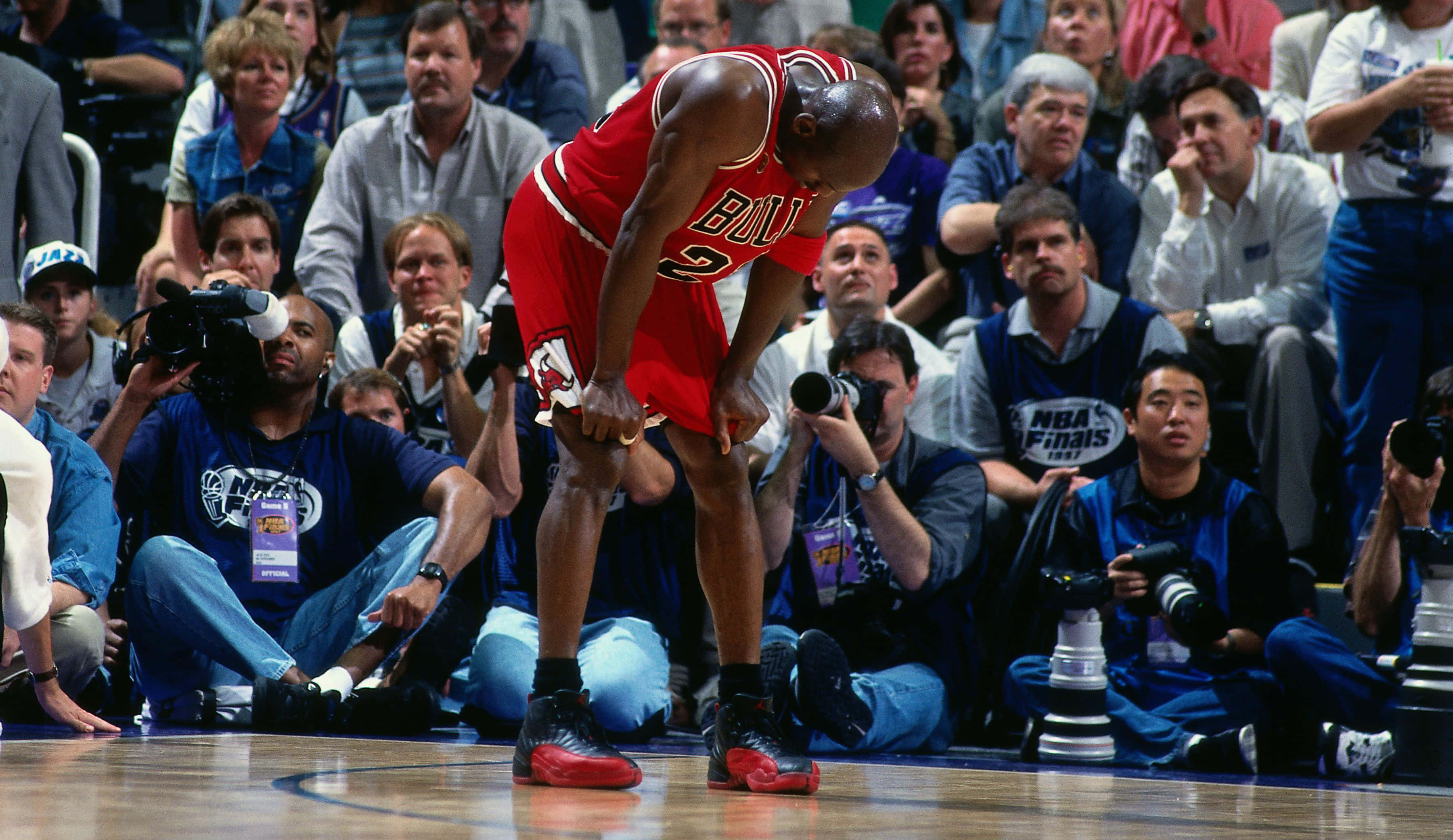 佐敦 桌布 壁紙 — 一位身穿紅色球衣的芝加哥公牛隊籃球運動員在NBA總決賽期間，在硬木球場上彎腰，周圍有攝影師和觀眾。