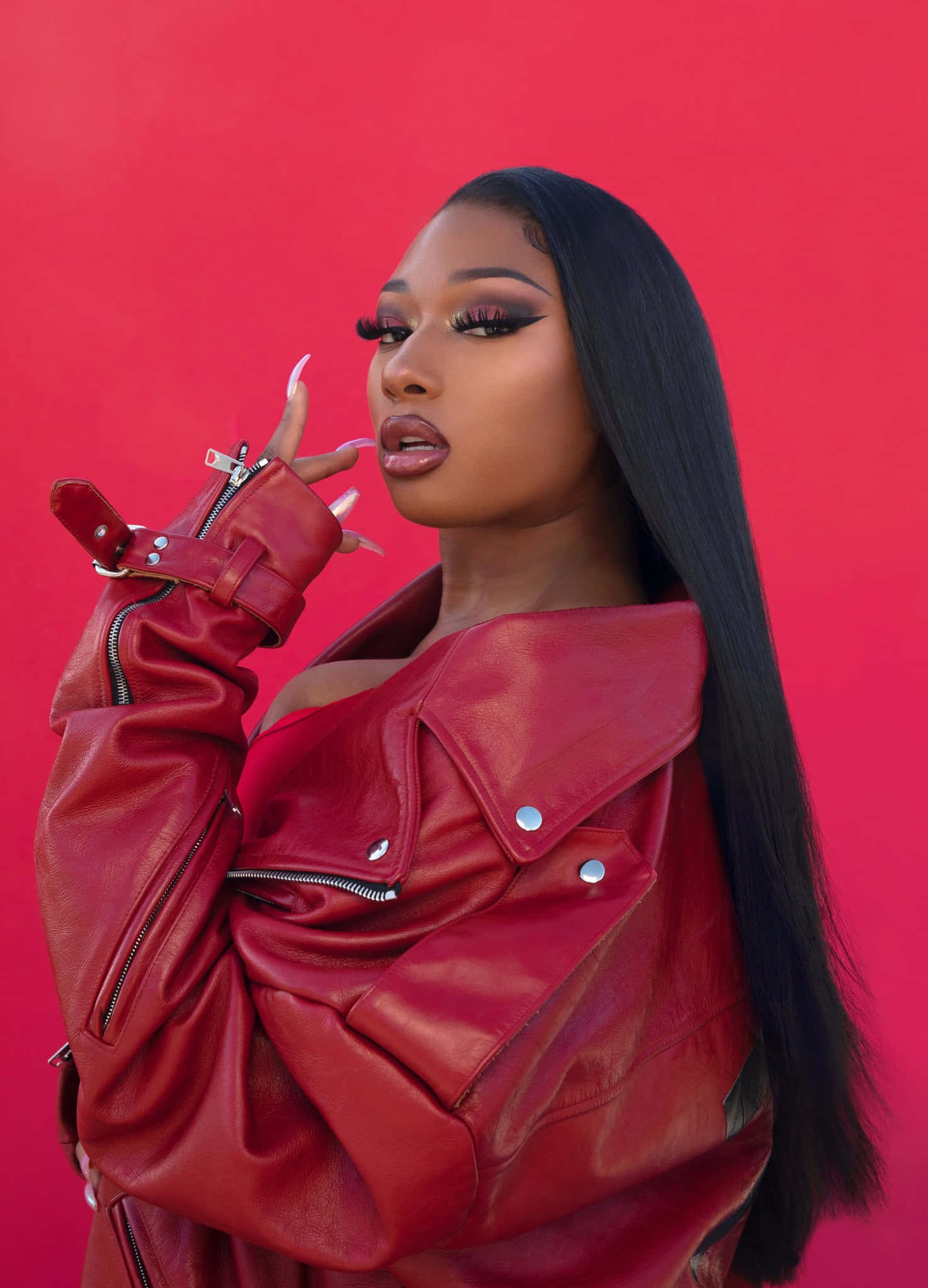 Megan Thee Stallion 桌布 壁紙 — 一位身穿紅色皮褸的女性，在純紅色背景前自信地擺姿勢，帶有戲劇性的化妝和長長的黑髮。