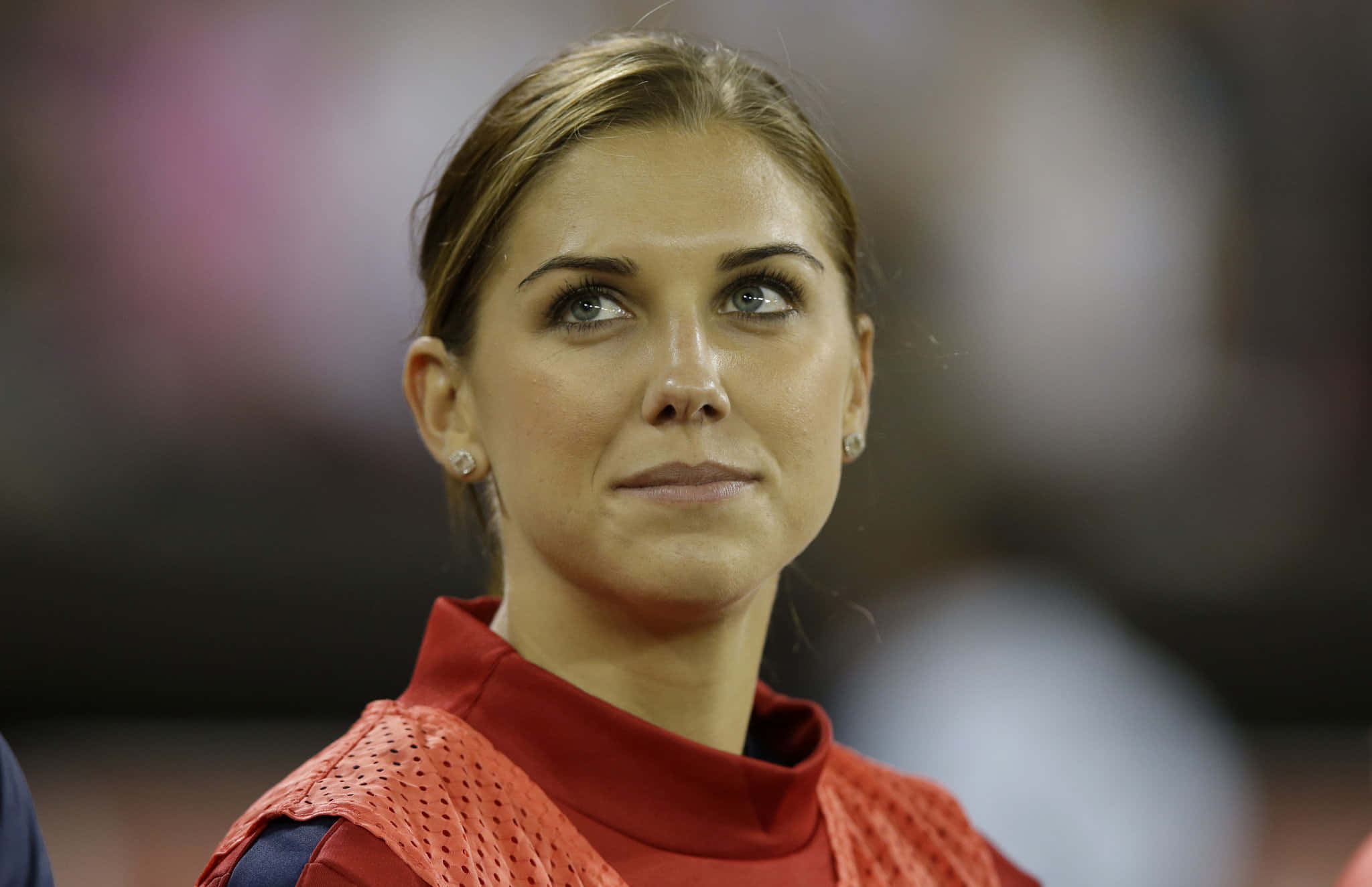 Alex Morgan 桌布 壁紙 — 一位身穿紅色穿孔上衣的女性，在模糊的體育場背景下，帶著沉思的表情向上凝視。
