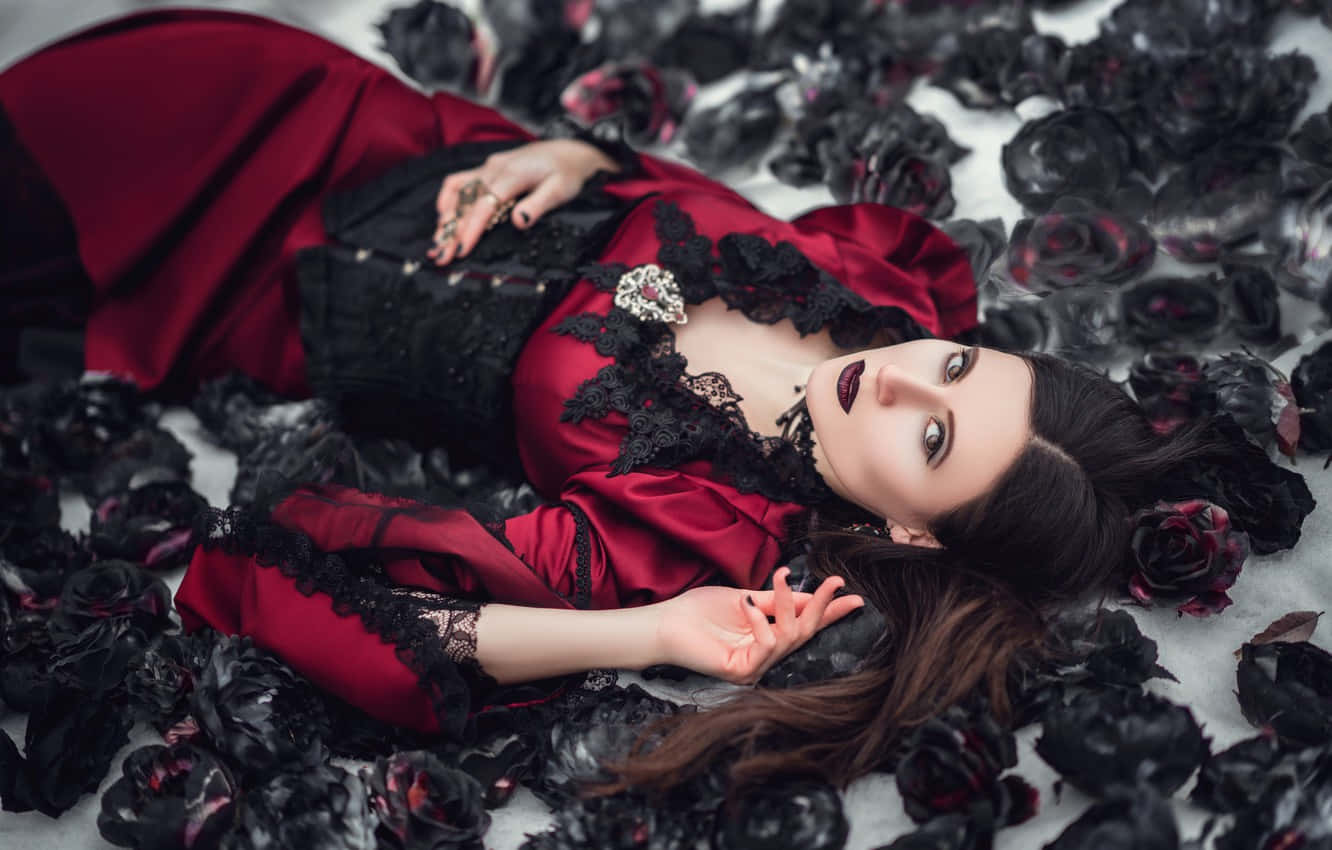 Gothic Fashion 桌布 壁紙 — 一位身穿紅色緞面禮服配黑色蕾絲的女性，躺在雪地上的一片黑玫瑰海中。