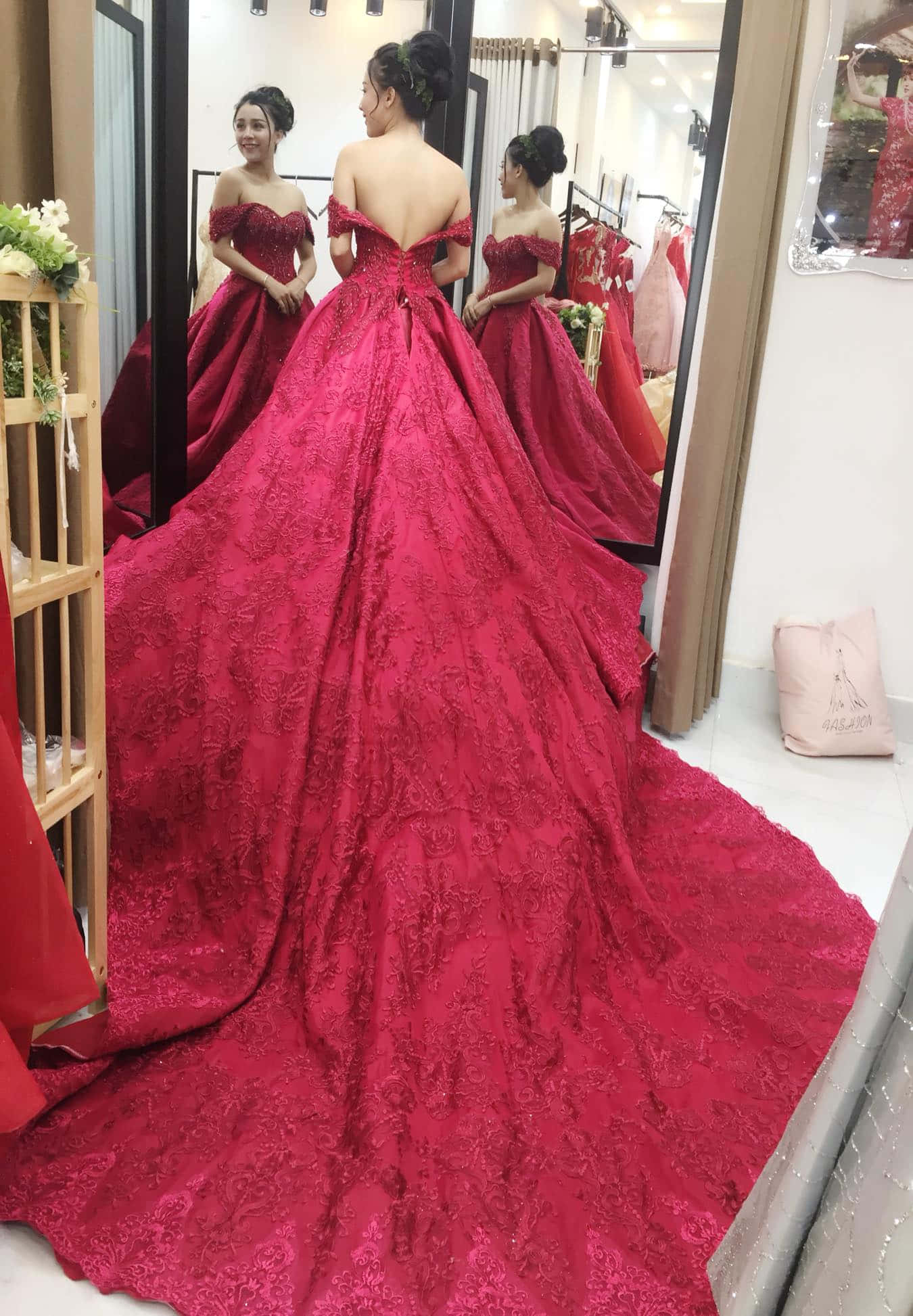 Red Wedding 桌布 壁紙 — 一位身穿紅色蕾絲婚紗的女子站在精品店的鏡子前，展示婚紗戲劇性的拖尾和露肩設計。