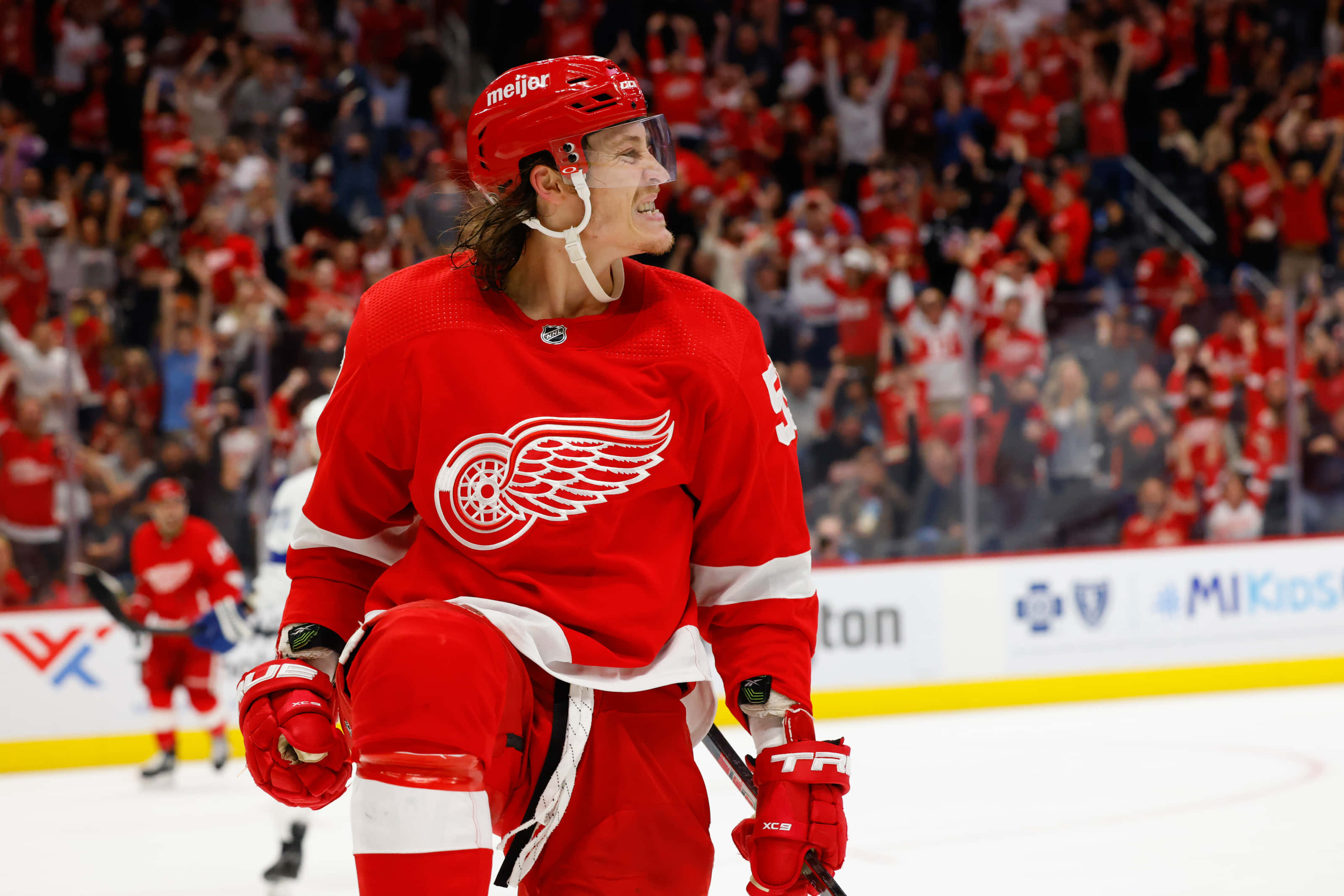 Tyler Bertuzzi 桌布 壁紙 — 一位身穿紅色裝備的底特律紅翼冰球運動員在冰上燦爛微笑，看台上的球迷熱情歡呼。