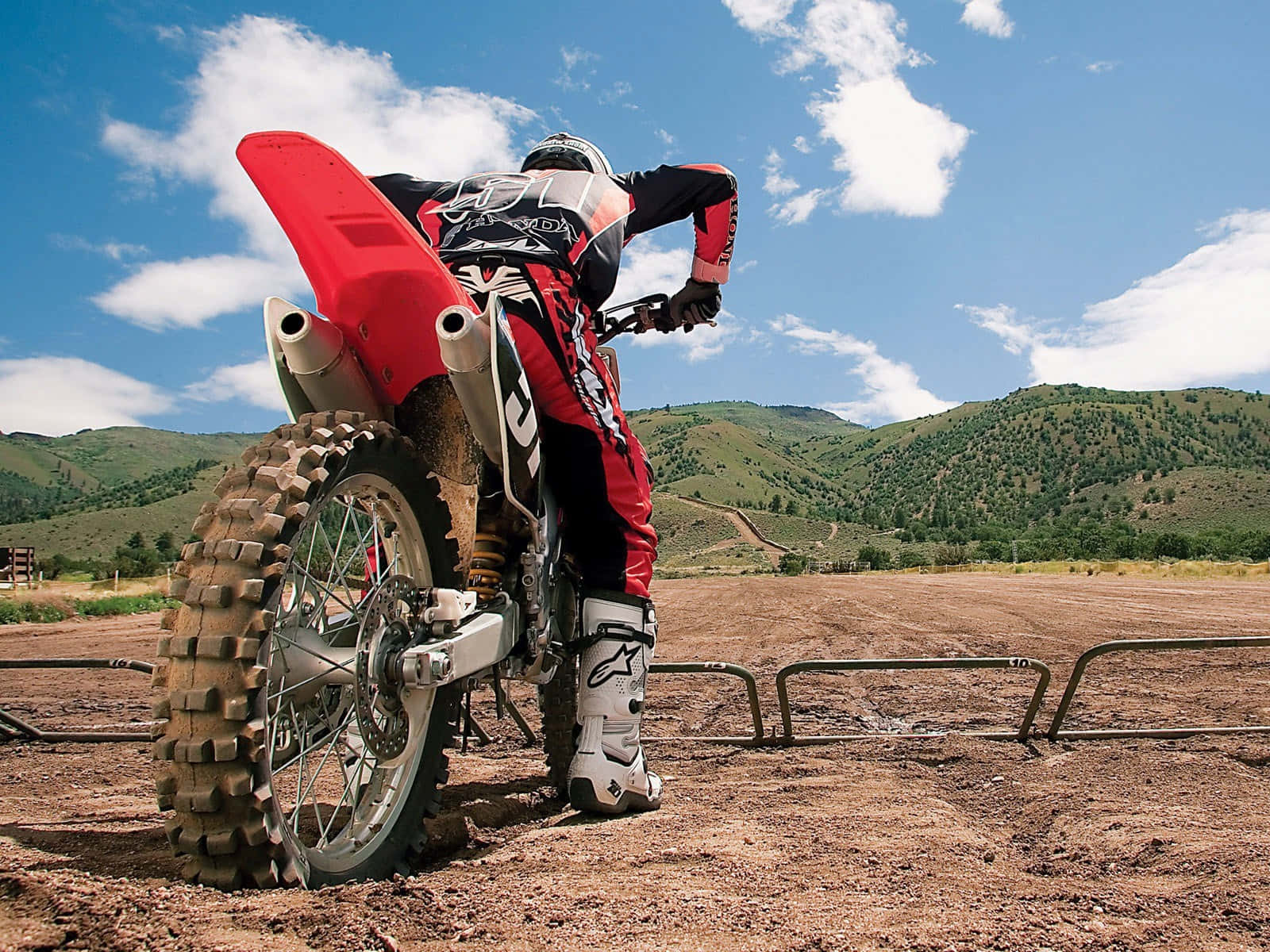 Motocross 桌布 壁紙 — 一位身穿紅色裝備的越野電單車手在泥地電單車上傾斜入彎，背景是起伏的綠色山丘和藍天。
