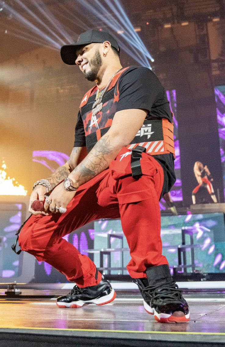 Anuel AA《Real Hasta La Muerte》 壁紙 — 一位身穿紅色褲子和黑色帽子的男性表演者蹲在舞台上，在明亮的舞台燈光下拿著麥克風。
