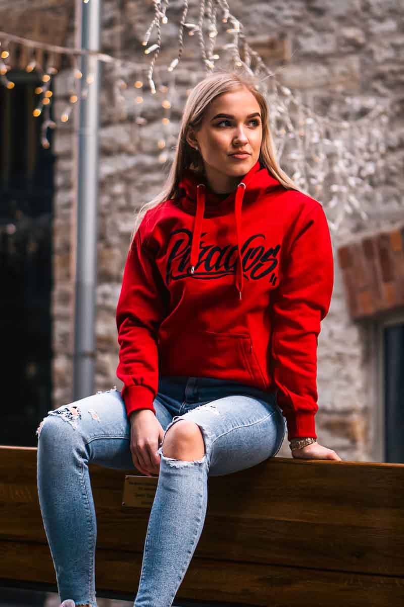 Red Hoodie 桌布 壁紙 — 一位身穿紅色連帽衫和破洞牛仔褲的年輕女性坐在靠著石牆的木長椅上，背景中串燈柔和地發光。