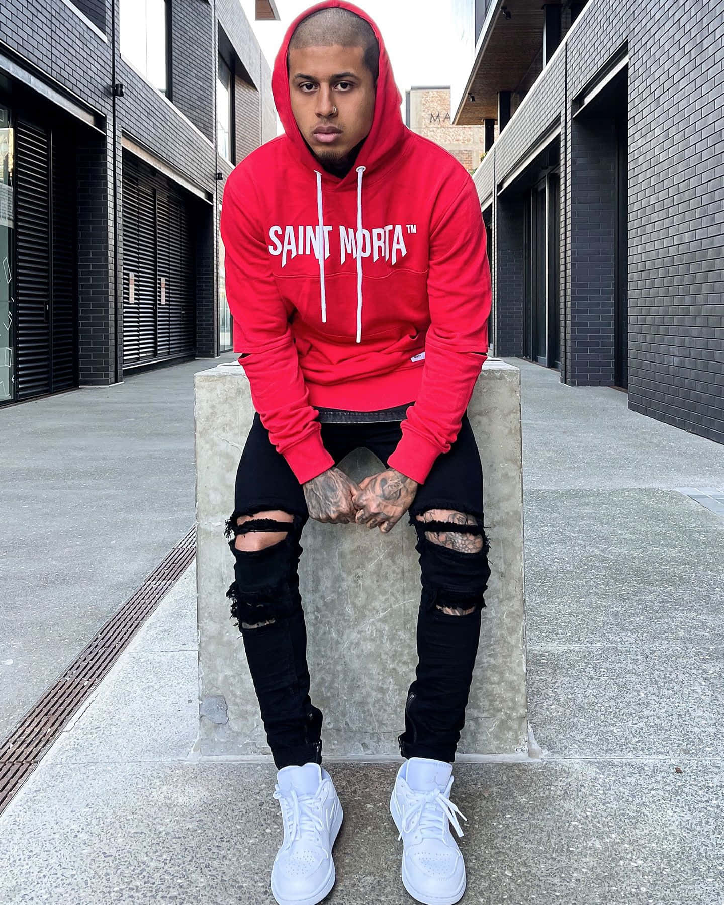 Red Hoodie 桌布 壁紙 — 一位身穿紅色連帽衫和破洞黑色牛仔褲的年輕男子自信地坐在現代城市小巷的混凝土塊上。