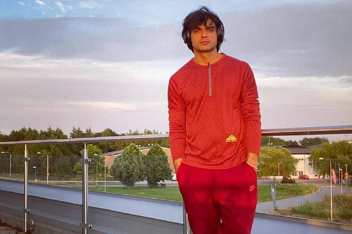 Neeraj Chopra 桌布 壁紙 — 一位身穿紅色運動服的年輕男子在黃昏時自信地站在現代陽台上，戴著耳機，凝視著柔和的天空。
