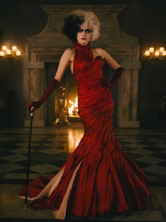 Cruella 2021 桌布 壁紙 — 一位身穿紅色長裙的引人注目的女性自信地站在宏偉、燈光昏暗的大宅中，身後是熊熊燃燒的壁爐。