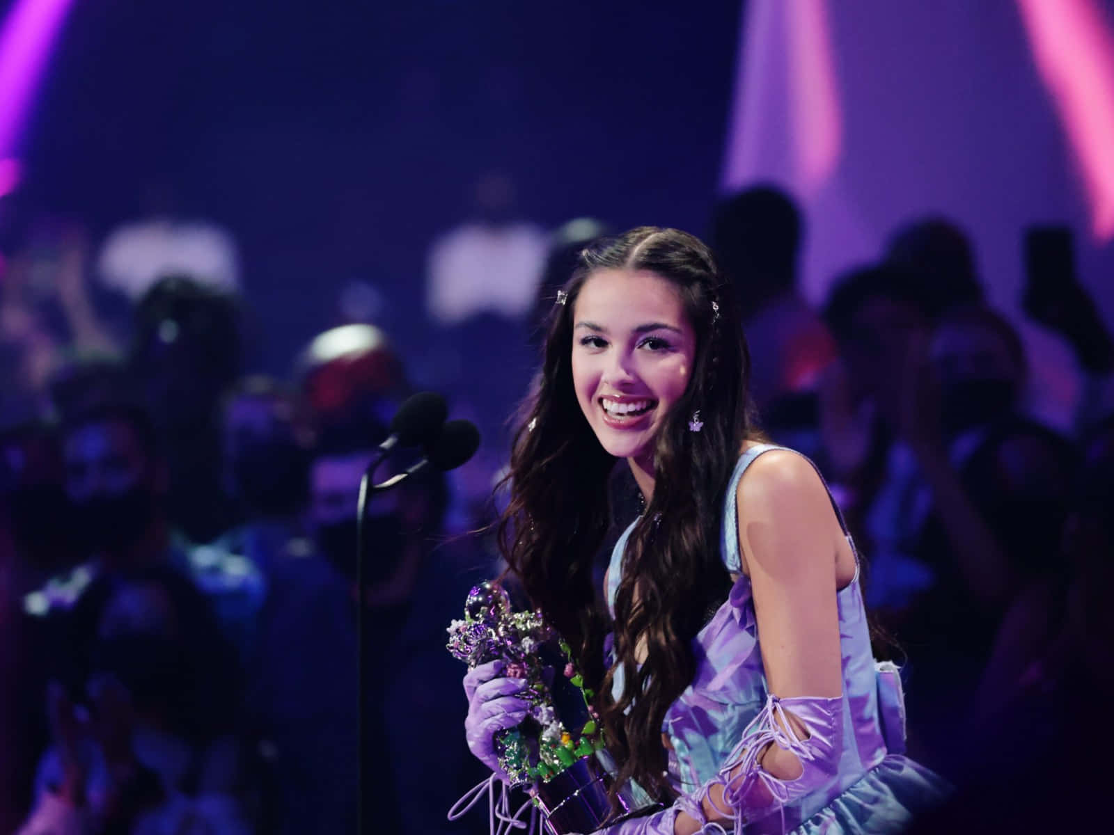 Olivia Rodrigo 桌布 壁紙 — 一位身穿紫色連衣裙的年輕女性在燈光璀璨的舞台上手持獎項，背後是模糊的觀眾，散發著光芒。