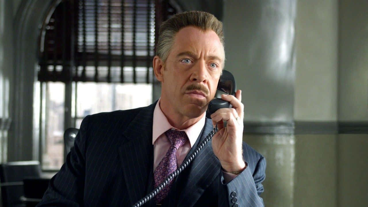 J Jonah Jameson 桌布 壁紙 — 一位身穿細條紋西裝的男子在現代辦公室裡拿著一部復古電話，表情嚴肅。