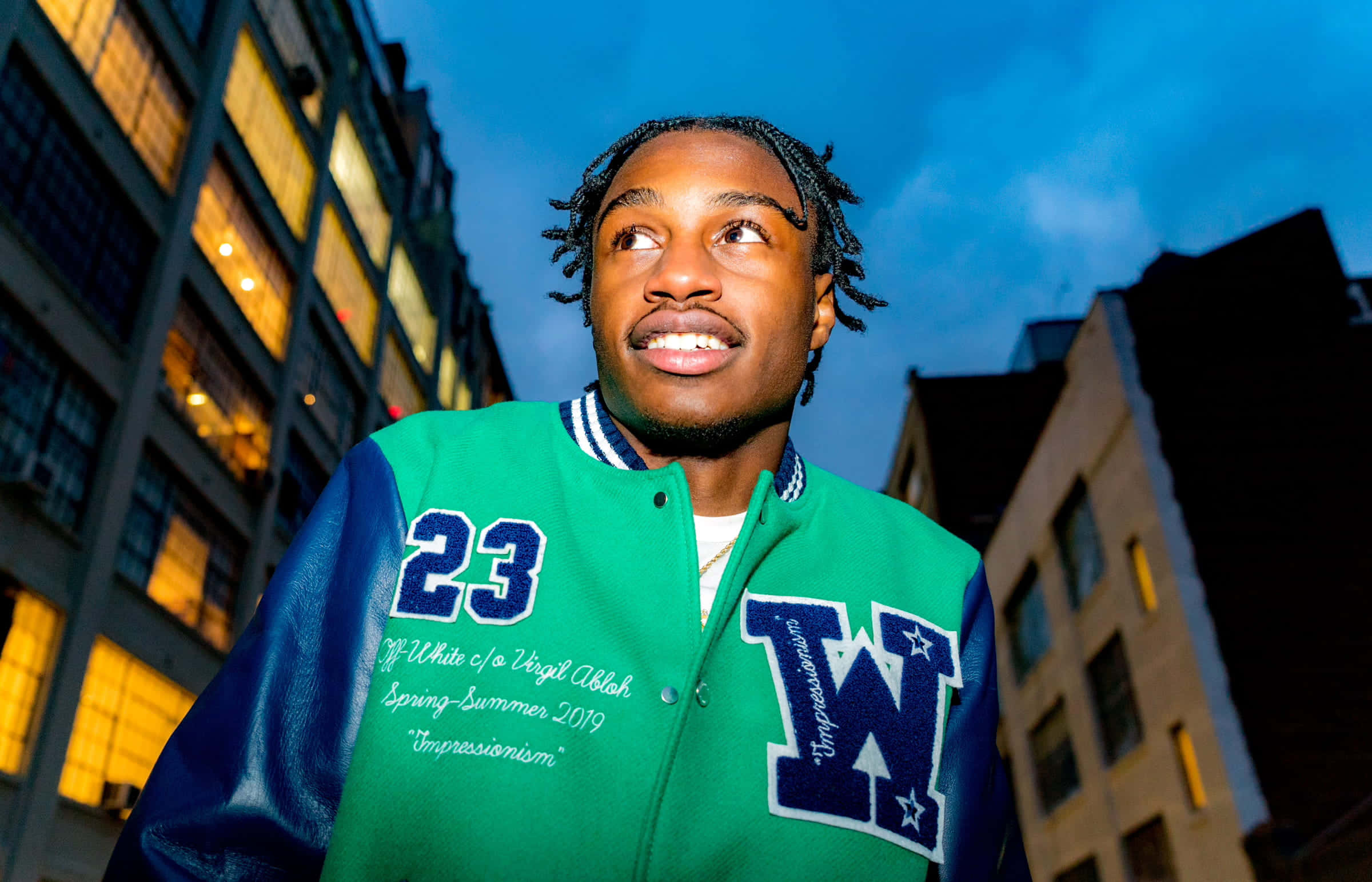 Lil Tjay 桌布 壁紙 — 一位身穿綠色和藍色Varsity外套的年輕男子，在暮色城市景觀中對著發光的窗戶向上微笑。