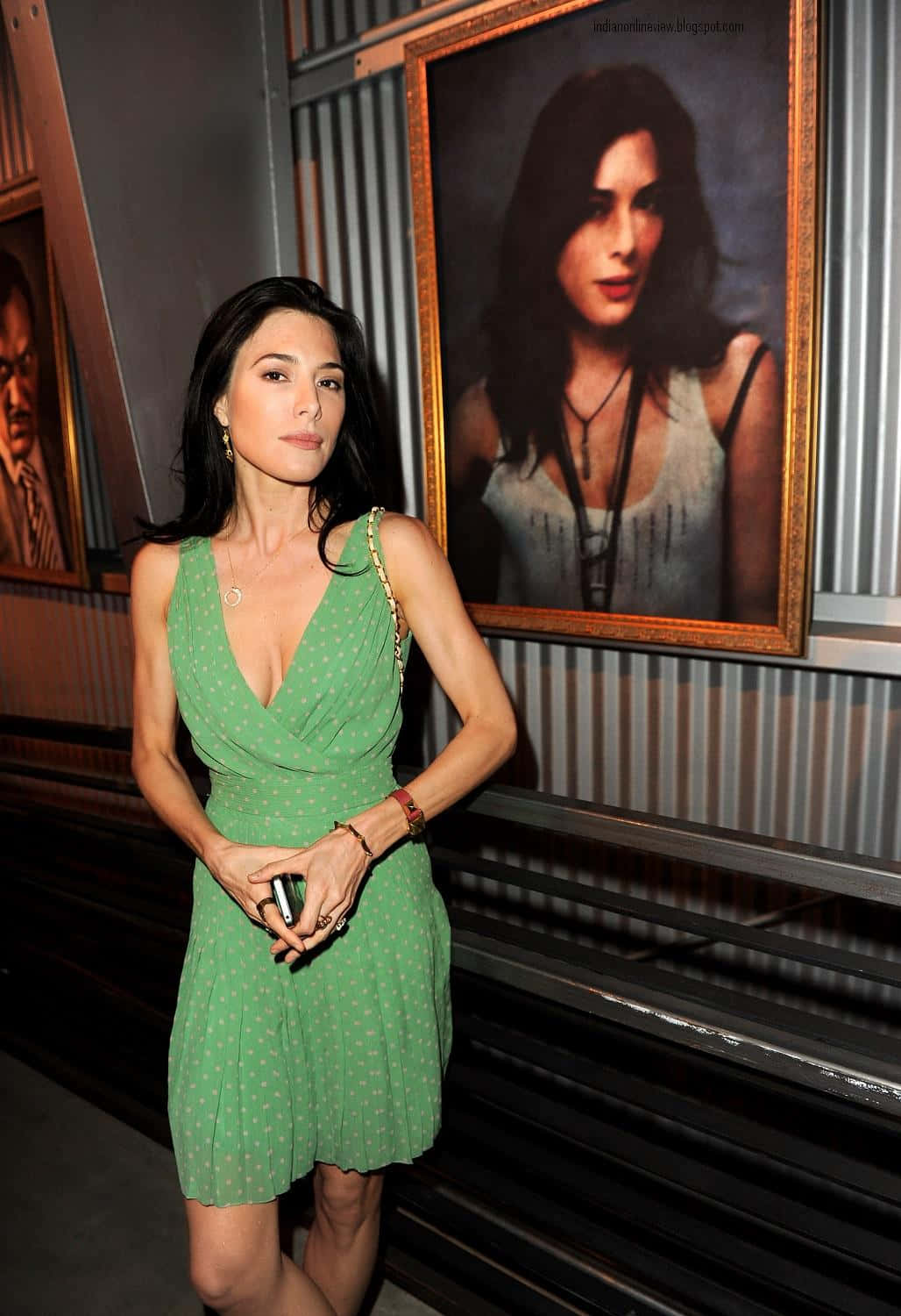 Jaime Murray 桌布 壁紙 — 一位身穿綠色圓點連衣裙的女性自信地站在一個光線昏暗、牆壁為波紋金屬的畫廊中，旁邊是鑲框肖像。