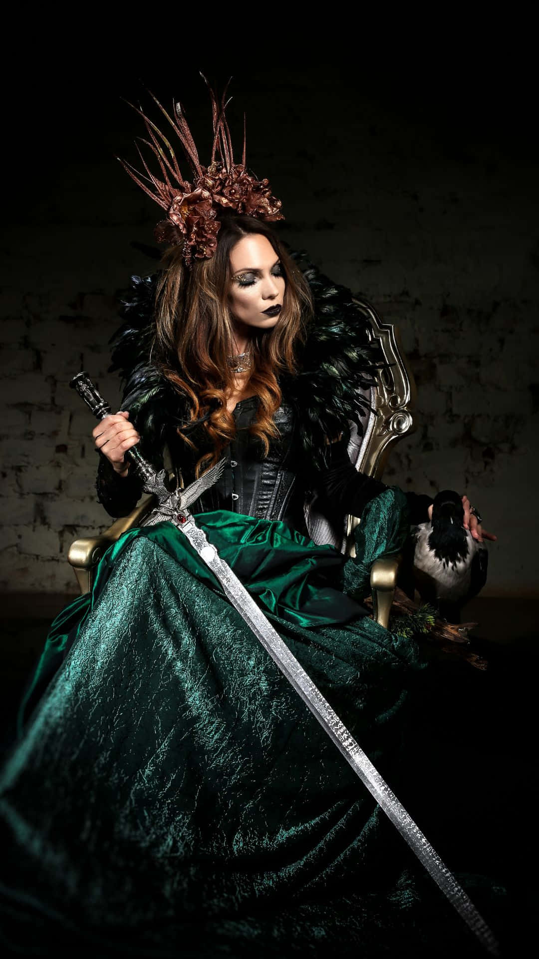 Horror Costumes 桌布 壁紙 — 一位身穿綠色天鵝絨長袍、頭戴尖刺王冠的黑暗女王威嚴地坐在王座上，手持一把劍，旁邊棲息著一隻烏鴉。