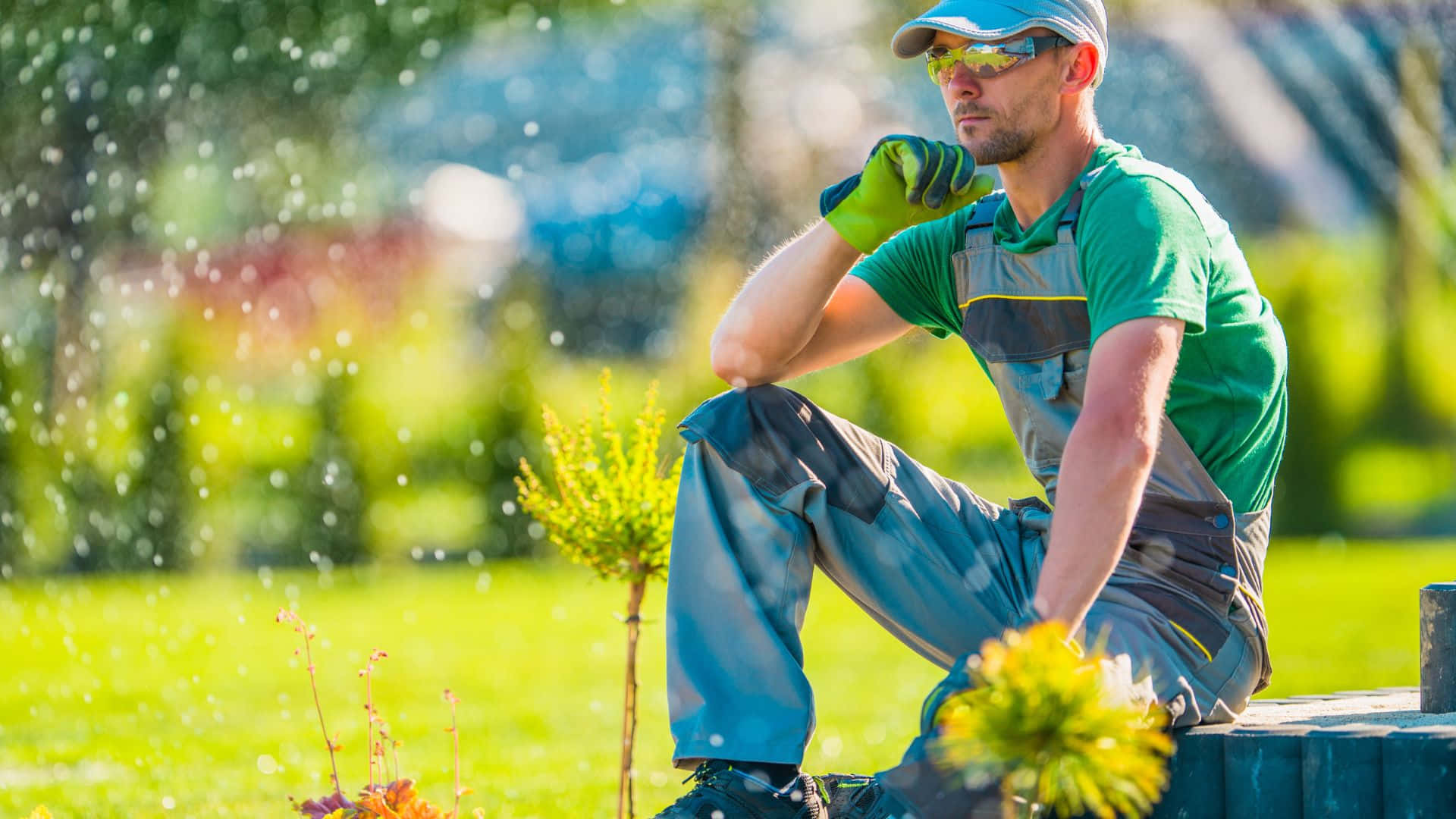 Garden Design 桌布 壁紙 — 一位身穿綠色工裝褲和手套的園丁坐在木長椅上，陽光普照的花園中水珠在他周圍噴灑。
