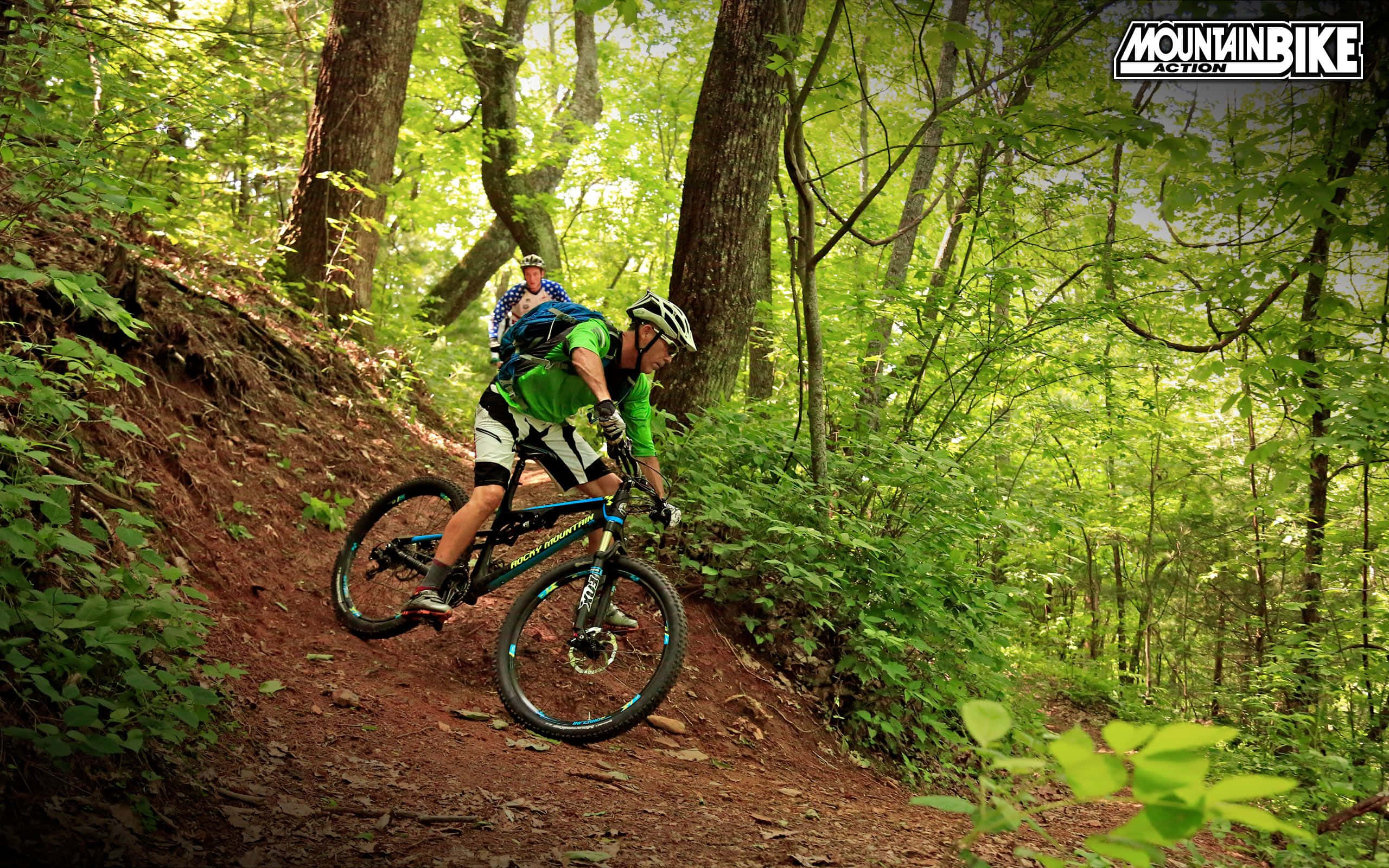 Mtb 桌布 壁紙 — 一位身穿綠色運動衫的山地自行車手在陽光明媚的森林小徑上，被茂密的綠樹和斑駁的光影包圍，傾身過彎。